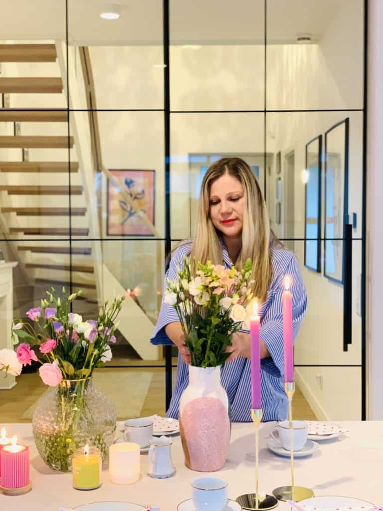 Aleksandra an ihrem Esstisch wie sie eine Blumenvase mit Schnittblumen bestückt. Im Hintergrund sieht man den Hausflur mit Treppe. 