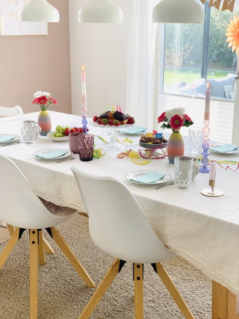 Esstisch aus Holz und Stühle mit weißen Sitzschalen und Holzbeinen im Scandi-Look. Der Tisch ist mit einer heller Tischdecke gedeckt und bunt für einen Geburtstag eingedeckt. 