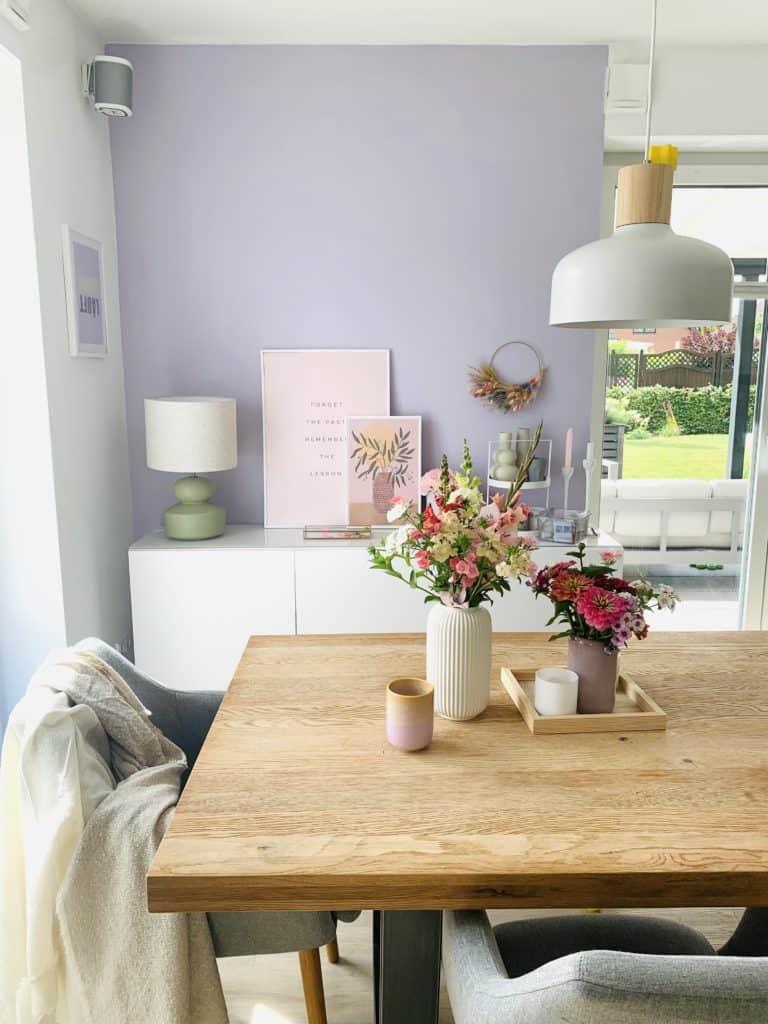 Esstisch aus Holz auf dem Blumenvasen stehen vor einem weißen Sideboard mit fliderfarbener Wand im Hintergrund  