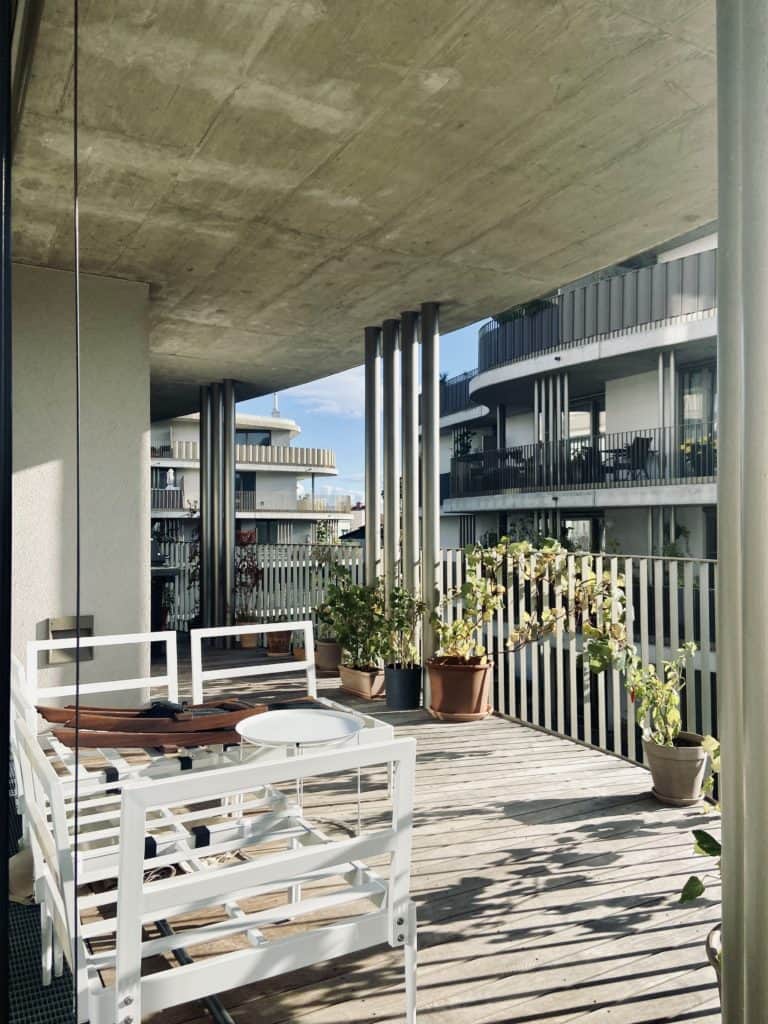 Funktional und familienfreundlich Wohnen: Großer Balkon mit Holzdielen und Sichtbeton-Decke mit weißer Sitzgruppe