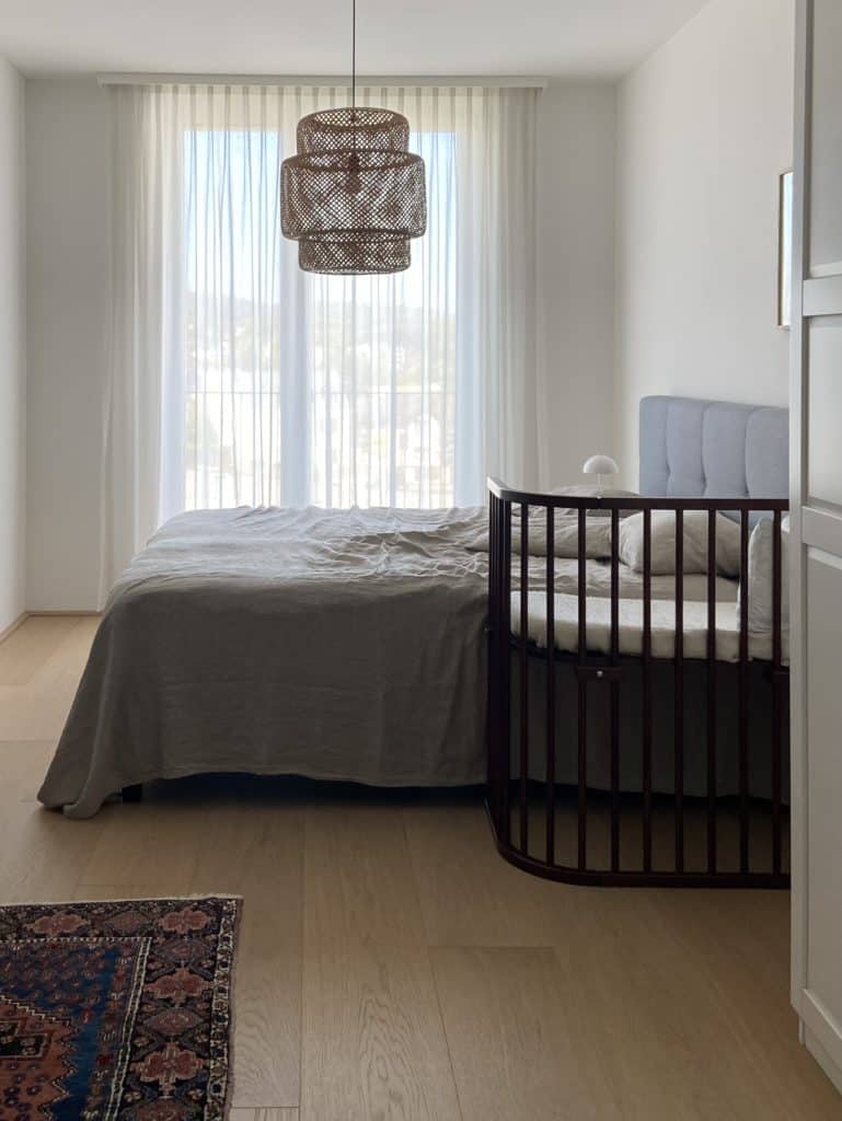 Minimalistisch eingerichtetes Schlafzimmer mit einem Doppelbett an dem ein Kinderbeistellbett steht. An der Decke hängt eine Deckenlampe aus Rattan.  Funktional und familienfreundlich Wohnen. 