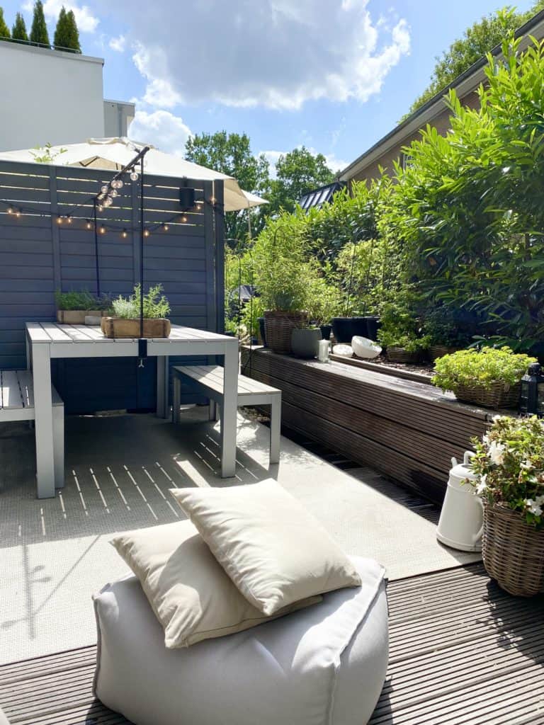 Soft Scandi und Industrial Minimalism - Großzügige Dachterrasse mit Bodenbelag aus Holzdielen mit Sitzbänken und Tisch aus Holz. Im Vordergrund befindet sich ein hellgrauer Outdoorsitzsack. 